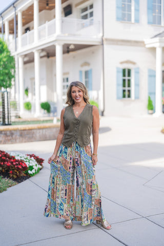 Woman posing outdoors in the Kaleidoscope Patchwork Flare Pants, a vibrant pair of wide-leg palazzo-style pants featuring an intricate patchwork micro-print in shades of turquoise blue, muted peach, sage green, and navy. Styled for a boho-chic look with an olive green button-front vest, metallic gold block-heel sandals, and gold jewelry in front of an elegant white building with light blue shutters.