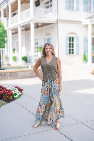 Full body front view of a smiling woman posing outdoors with one hand on her hip, modeling a tailored olive green button-front vest. The waistcoat-style top features a classic V-neckline, faux front welt pockets, and tonal covered buttons. She pairs the sleeveless vest with flowy, wide-leg pants featuring a vibrant, multi-colored patchwork print filled with intricate floral and geometric patterns. The bohemian-chic aesthetic is completed with metallic gold open-toe block heels, a gold pendant necklace, and 