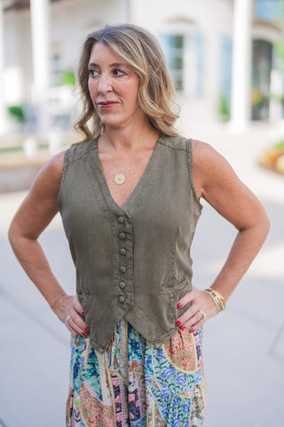 Front portrait view of a woman posing outdoors with hands on her hips, modeling a tailored olive green button-front vest. The waistcoat-style top features a classic V-neckline, tonal covered buttons, faux front welt pockets, and a pointed hem. The sleeveless vest is paired with vibrant, multi-colored patchwork print pants showcasing intricate floral and geometric patterns. She completes the bohemian-chic aesthetic with a gold compass pendant necklace, gold bangles, and a bold red manicure, set against a cla