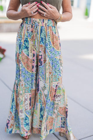 A close-up view of the Kaleidoscope Patchwork Flare Pants highlighting the elasticized high waist with an adjustable drawstring tie. Demonstrates the fluid, lightweight fabric drape and tiered flared leg construction, showing off the detailed combination of floral, geometric, and paisley patchwork motifs alongside metallic gold open-toe heels.