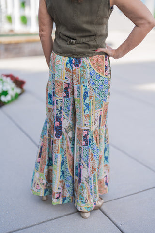 Back view of the Kaleidoscope Patchwork Flare Pants showcasing the seamless flow of the multi-colored patchwork print across the seat and full, voluminous legs. Highlights the comfortable stretch waistband and dramatic tiered flare silhouette styled with an olive vest tailored back-buckle detail outdoors.
