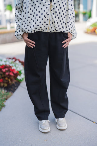 Lower body front view of a woman modeling black barrel-leg pants featuring distinct, architectural seam detailing down the front of the legs. The relaxed-fit, structured trousers feature comfortable front slant pockets where the model is resting her hands. The pants are styled for a chic everyday look with a white and black polka dot zip-up jacket and white platform sneakers with metallic gold accents, photographed outdoors on a paved walkway.