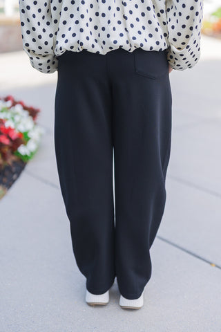 Lower body back view of the black barrel-leg pants highlighting their comfortable, wide-leg drape and smooth fit. The tailored back features a clean waistband and a single back pocket on the right side. The pants fall effortlessly over the white platform sneakers, paired perfectly with the playful polka dot top for a stylish, transitional outfit.