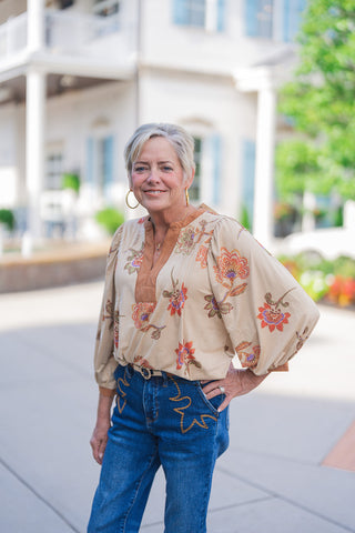 Woman smiling outdoors wearing a beige vintage-inspired floral blouse featuring voluminous balloon sleeves and a contrasting tan faux-suede trim around a split V-neckline. The relaxed-fit top is casually front-tucked into medium-wash blue denim jeans that showcase unique western-style star embroidery on the front pockets. Styled with a tan belt and large gold hoop earrings, she is standing on a paved walkway in front of a modern white building with light blue shutters.