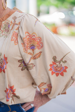 A detailed close-up view of the beige floral blouse highlighting the gathered pleating at the shoulder that creates the architectural, dramatic balloon sleeve. The image emphasizes the rich, warm-toned botanical print—featuring intricate orange, rust, purple, and olive green florals—alongside the soft, contrasting tan trim finishing the cuffs and neckline.