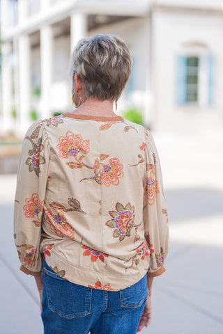 Back view of the beige floral blouse showcasing the relaxed, comfortable drape of the fabric and a clean central vertical seam. The large-scale, vibrant floral pattern spans the back gracefully, framed by the continuous contrasting tan trim along the back of the neck and the bottom hemline, paired effortlessly with classic blue jeans.