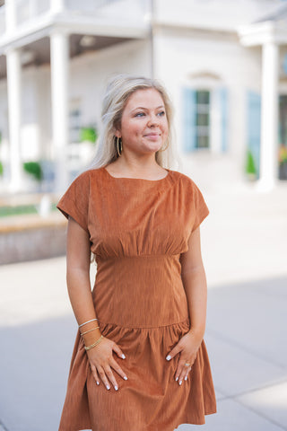 A portrait view of the rust mini dress highlighting the rich, crinkled and ribbed texture of the fabric. The design features a subtly gathered bodice that seamlessly meets a wide, structured waistband, creating a flattering and comfortable silhouette. The model looks off-camera, accessorized with gold hoop earrings and delicate layered gold bracelets.
