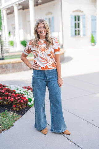 Full body front view of a smiling blonde woman posing outdoors with one hand on her hip, modeling a short-sleeve cream knit top featuring a vibrant orange and tan tropical botanical pattern with palm leaves and floral motifs. The fitted sweater top features ribbed banding at the crew neckline, sleeves, and hem. She pairs it with medium-wash blue denim wide-leg jeans detailed with vertical side seams. The casual, warm-weather look is completed with tan suede pointed-toe shoes, a gold chain necklace, and gold