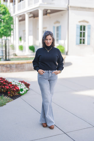 Full body front view of a woman posing outdoors with her hands resting in her front pockets, modeling a cozy black ribbed turtleneck sweater. The knit top features long balloon sleeves with fitted cuffs and a comfortable, slightly cropped fit that sits perfectly at the waistline. She pairs the sweater with light grey wash denim jeans featuring a subtle flare-leg silhouette. The chic, transitional fall outfit is accessorized with a gold cross pendant necklace, gold hoop earrings, a gold watch, and brown clog