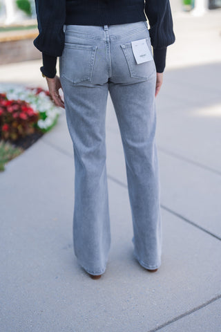 Lower body back view focusing on the tailored, flattering fit of the light grey wash denim jeans. The image highlights the smooth drape through the hips, classic back patch pockets, and a subtle flare hem that rests effortlessly over brown leather shoes. A visible white Vervet brand tag is attached to the right back pocket, pairing seamlessly with the black ribbed sweater for a trendy, texture-rich everyday aesthetic.