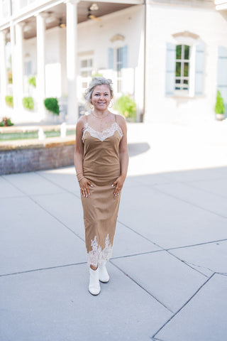 Woman posing outdoors in the Camel Faux Suede Lace-Trim Slip Dress, an elegant tan midi dress featuring a soft, suede-like texture. The garment boasts a delicate cream eyelash lace V-neckline and a matching wide scalloped lace hem. Styled with chic white ankle boots and dainty gold jewelry, standing on a paved courtyard in front of a classic white building with columns and light blue shutters.