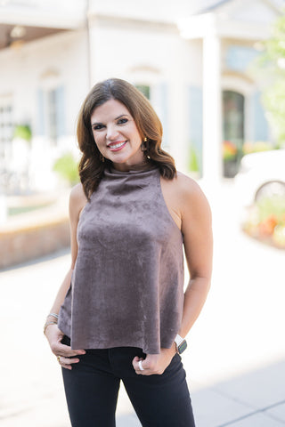Woman posing outdoors in the Taupe Faux Suede Halter Top, a chic sleeveless piece featuring a soft, velvety texture and a sophisticated high mock neckline. The relaxed-fit swing bodice drapes elegantly, offering a comfortable yet elevated silhouette. She is styled casually with black denim jeans, geometric drop earrings, stacked bracelets, and a white smartwatch, standing smiling with her hands in her pockets in front of an elegant white building with classical columns and light blue shutters.
