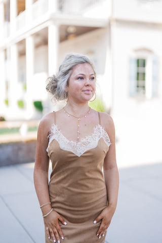 A portrait view of the Camel Faux Suede Lace-Trim Slip Dress highlighting the romantic, intricate details of the bodice. The soft tan faux suede fabric contrasts beautifully with the sheer cream eyelash lace outlining the plunge V-neckline. The dress features thin spaghetti straps and is accessorized with gold hoop earrings and a delicate pearl-accented gold Y-drop necklace, elevating the effortless, romantic aesthetic.
