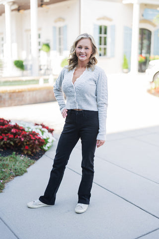 Woman posing outdoors in the Grey Lace-Trim Cropped Cardigan, a cozy light grey sweater featuring a V-neckline accented with delicate white scalloped lace. The long-sleeve knit top showcases raised bobble stitching, a classic three-button front closure, and a ribbed cropped hem. She is styled casually in high-waisted black jeans and glittery silver sneakers, standing on a paved walkway in front of an elegant white building with classical columns and light blue shutters.