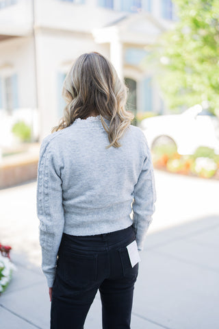 Back view of the Grey Lace-Trim Cropped Cardigan showcasing the smooth, uninterrupted knit pattern across the back which contrasts elegantly with the textured sleeves. The image highlights the wide, ribbed banded hem resting neatly at the natural waistline of the high-rise black denim jeans, finished with a subtle peek of the white lace trim at the nape of the neck.
