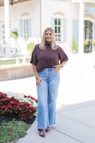 Woman posing outdoors in a fall-ready outfit featuring a deep burgundy ribbed knit top with short dolman sleeves, partially front-tucked into high-waisted, light-wash wide-leg jeans. The transitional seasonal look is completed with striking burgundy western boots featuring white embroidery, and accessorized with stacked gold, pearl, and red bangle bracelets. She stands confidently on a paved walkway next to a vibrant flower bed, with an elegant white building featuring classical columns and light blue shutt