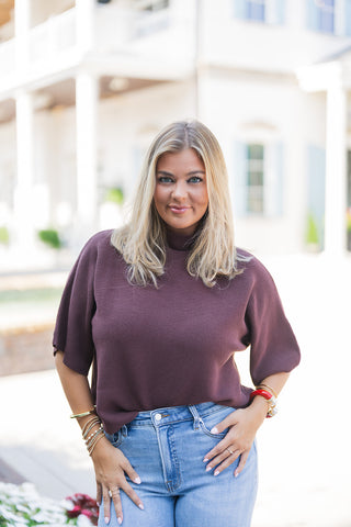 A portrait view of a woman wearing the Burgundy Ribbed Dolman Top, highlighting the rich, autumnal hue and fine horizontal ribbed texture of the knit fabric. The relaxed-fit sweater features a sophisticated high mock neckline and breezy short dolman sleeves. Casually front-tucked into high-waisted, light-wash blue jeans, the look is styled with a stack of gold, beaded, and bold red bangle bracelets. She stands smiling in front of an elegant white building with classical columns and light blue shutters out o