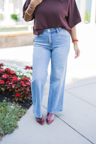 A close-up front view focusing on the tailored fit of the high-waisted, light-wash wide-leg denim and the tucked-in burgundy ribbed knit top. The image highlights the classic denim front button closure and pocket styling, as well as the intricate white stitching on the pointed toe of the burgundy cowboy boots peeking out from beneath the wide hem. The model's hands rest at her sides, showcasing layered gold and red bracelets that perfectly tie the casual, elevated western aesthetic together.
