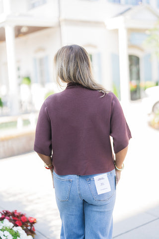 Back view of the Burgundy Ribbed Dolman Top showcasing the clean, uninterrupted drape of the horizontal ribbed knit. The image highlights the relaxed, slightly boxy silhouette and the comfortable short sleeves as the straight, untucked back hem falls effortlessly over the waistband of classic light-wash denim jeans featuring standard back pockets.