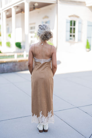 Back view of the Camel Faux Suede Lace-Trim Slip Dress showcasing the alluring low scooped back design, accented with a horizontal panel of cream lace trim. The image highlights the adjustable spaghetti straps, the smooth, uninterrupted drape of the faux suede fabric down the silhouette, and the striking lace detailing along the bottom hem, finished perfectly with classic white leather ankle boots.