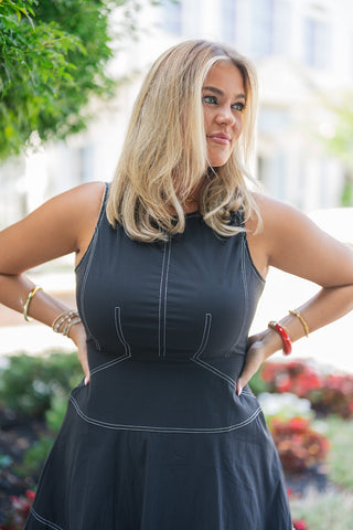 A portrait view of the Black Contrast Stitch Drop Waist Mini Dress highlighting the architectural detail of the crisp white topstitching against the smooth black fabric. The close-up emphasizes the flattering corset-like curved princess seams on the bodice, the horizontal drop-waist seam, and the classic high neckline. The sleeveless cut provides a breathable, tailored fit, perfectly accessorized with a bold red bangle and stacked gold bracelets.