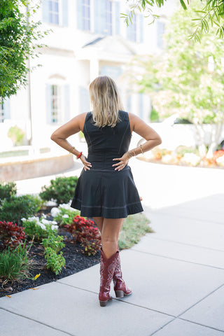 Back view of the Black Contrast Stitch Drop Waist Mini Dress showcasing the continuous, contoured white topstitching that runs down the center back and elegantly traces the drop-waist silhouette. The image highlights the full back coverage of the sleeveless bodice and the fluid, airy flare of the mini skirt's hemline. The look is grounded by the striking burgundy western boots, offering a complete view of the outfit's flattering proportions and styling.