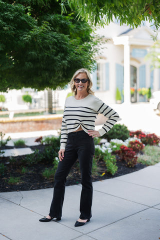 Woman posing outdoors in the Breton Stripe Twist-Front Sweater, a chic cream and black horizontally striped long-sleeve knit top featuring a classic crew neckline and a unique twisted knot detail at the front hem. The slightly cropped, asymmetrical silhouette is effortlessly styled with high-waisted black jeans, black pointed-toe flat shoes, and dark sunglasses. She is standing on a paved walkway in a bright outdoor setting with manicured greenery and an elegant white building in the background.