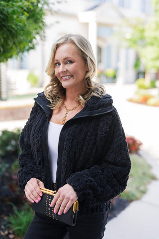 A close-up portrait view highlighting the textured pattern of the Textured Fleece Zip-Up Jacket in black. The design features a plush, tactile weave, structured stand collar, and central front zipper. Styled over a white top with a gold chain necklace and a black studded clutch wallet, offering a chic, elevated look for cool-weather layer styling.