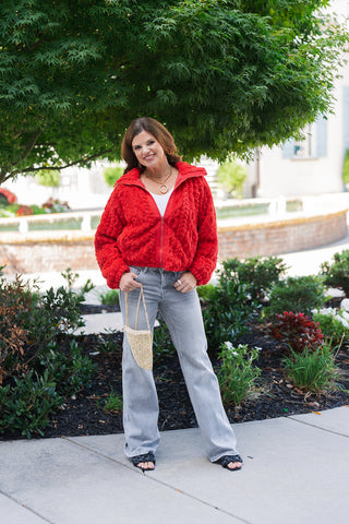 Woman posing in the Textured Fleece Zip-Up Jacket in vibrant red, a cozy statement layer featuring a chunky cable-knit textured fleece design, high collar, and front zip closure. The relaxed jacket is styled over a white top and paired with light wash grey wide-leg jeans, black woven slide sandals, gold chain necklaces, and a small woven shoulder bag outdoors under a leafy green tree.