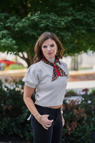 A portrait view of the Short Puff Sleeve Mock Neck Sweater in light beige, highlighting the smooth knit texture and tailored bodice. The close-up emphasizes the gathered shoulder seams that create the structured puff sleeves and the high mock neck collar, perfectly framed by the tied leopard print scarf. Styled casually with black denim, geometric tortoiseshell earrings, and stacked metallic bracelets.