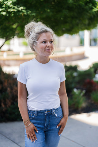 Woman looking off-camera outdoors wearing the cotton Good American baby tee. The image highlights the smooth, tailored fit of the short-sleeve tee and its classic, structured crew neckline, resting cleanly at the waistline of medium-wash blue jeans. Accessorized with gold hoop earrings and layered delicate bracelets, showcasing a highly versatile, staple wardrobe aesthetic.