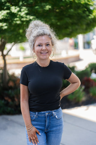 Woman smiling outdoors wearing the cotton Good American baby tee in black. The classic short-sleeve top features a high crew neckline and a flattering, form-fitting silhouette that smooths the torso. Styled casually for an effortless, everyday look with medium-wash blue jeans, large gold hoop earrings, a delicate gold necklace, and stacked bracelets. The background features a bright, blurred courtyard with a lush green tree and a paved walkway.