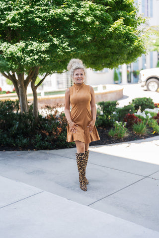 Woman posing outdoors in the Camel Faux Suede Mock Neck Mini Dress, a chic sleeveless tan dress featuring a structured high neckline and vertical princess seams for a tailored bodice. The dress finishes above the knee with a playful, fluted ruffle hem. She is styled boldly with statement knee-high leopard print block-heel boots and large gold hoop earrings, standing on a paved walkway in front of a lush green tree and an elegant courtyard.