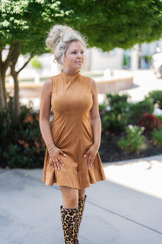 A portrait view of the Camel Faux Suede Mock Neck Mini Dress highlighting the architectural detail of the high mock neckline and the clean, sleeveless shoulder cut. The soft, camel-colored faux suede fabric offers a rich, velvety texture, complemented by the precise vertical topstitching that contours the waistline. Styled with a relaxed high bun, gold hoop earrings, and delicate stacked bracelets for a trendy, transitional seasonal aesthetic.