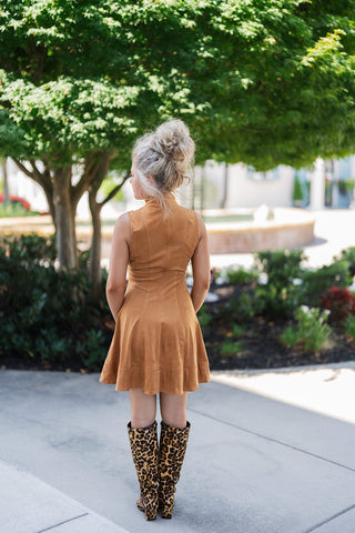 Back view of the Camel Faux Suede Mock Neck Mini Dress showcasing the continuous vertical tailored seams running down the back to create a flattering, contoured silhouette. The image highlights the full back coverage of the high neckline, the smooth faux suede drape, and the energetic movement of the flared drop-hem. The striking knee-high leopard print boots perfectly anchor the autumnal look from behind.