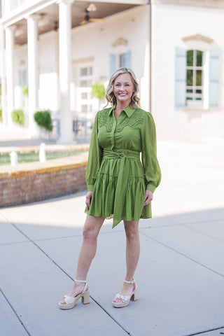 Woman smiling and posing in a vibrant olive green long-sleeve mini dress featuring an intricate eyelet crochet lace yoke across the chest and shoulders. The dress highlights a classic pointed collar, a button-down front bodice, long bishop sleeves with fitted cuffs, and a self-tying fabric waist belt with a ring buckle. The skirt falls into a flared, tiered ruffled hemline. Styled for a chic daytime aesthetic with beige espadrille platform sandals, photographed outdoors on a sunny walkway in front of a whit