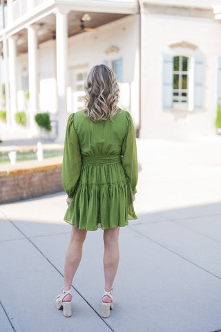 Back view of the olive green mini dress showcasing the gathered details at the back waistline and the flowy, full movement of the tiered ruffled skirt. The image highlights the light, textured fabric drape across the back and shoulders, the long bishop sleeves, and the wide tie-belt cinching the waist, offering a complete look at this romantic, feminine silhouette paired with woven platform heel sandals.