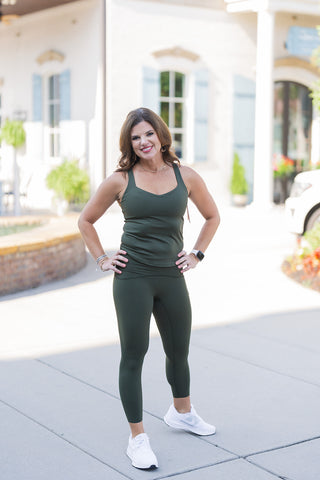 Woman posing outdoors in a matching olive green activewear set, featuring a fitted V-neck tank top with delicate shoulder straps and high-waisted, ankle-length leggings. The smooth, form-fitting fabric creates a sleek and supportive silhouette. Styled for an everyday athleisure aesthetic with bright white running sneakers and layered metallic bracelets, photographed on a sunny paved walkway in front of a classic white building with light blue shutters.
