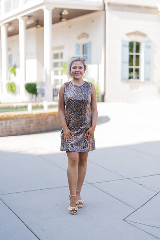 Woman posing outdoors in a glamorous sleeveless mini dress featuring a classic brown and black leopard print overlaid with shimmering vertical sequin stripes. The dress has a structured high crew neckline finished with delicate black contrast piping. Styled for a chic, party-ready aesthetic with metallic gold ankle-strap espadrille wedges featuring a knotted toe detail, and large gold hoop earrings. She is standing on a paved walkway in front of an elegant white building with classical columns and light blu