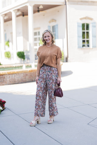 Woman smiling and posing outdoors in a chic, transitional outfit featuring a short-sleeve camel-brown knit sweater with a central cable-knit detail. The top is partially tucked into high-waisted, wide-leg pants showcasing an intricate botanical paisley print in earthy shades of teal, brown, burgundy, and cream. Styled for a polished everyday aesthetic with beige espadrille platform sandals and a burgundy pouch handbag with a gold ring handle, photographed on a sunny paved walkway in front of a classic white
