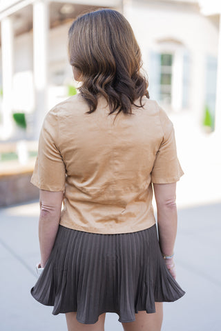 Back view of the camel faux suede top and olive-brown pleated skirt, showcasing the garment's continuous tailored details. The top features a sharp vertical center seam intersected by a horizontal mid-back stitch, providing a structured yet relaxed fit. The short-sleeve top drapes cleanly at the waistline, perfectly complementing the crisp, uniform folds of the pleated mini skirt for a balanced, sophisticated silhouette.