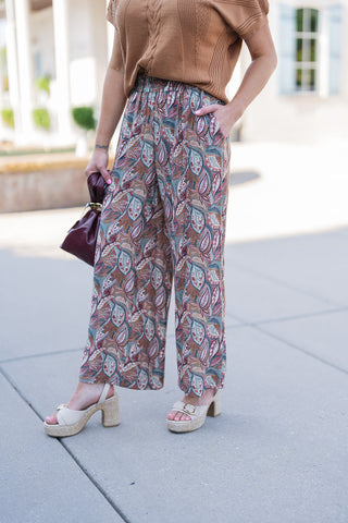 A mid-body and leg close-up focusing on the tailored yet relaxed fit of the botanical paisley wide-leg pants. The image highlights the comfortable, smocked elastic waistband and a functional side pocket where the model's hand is casually resting. The flowy, cropped wide-leg silhouette falls gracefully over beige espadrille platform sandals, while the rich texture of the camel knit sweater and the burgundy handbag add elevated, seasonal contrast.