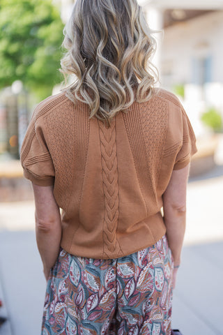 Back view of the Caramel Macchiato Knit sweater, showcasing the continuous central cable knit braid running down the spine, flanked by structural ribbed panels and subtle pointelle detailing. The short, slightly puffed sleeves and softly banded hem provide a clean, tailored silhouette that drapes elegantly over the paired patterned wide-leg pants.