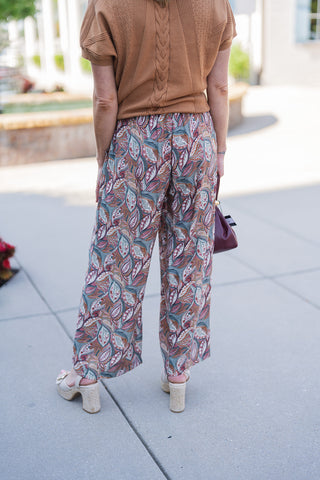 Back view of the outfit showcasing the relaxed drape of the short-sleeve camel knit sweater over the patterned wide-leg pants. This angle emphasizes the fluid, airy movement of the lightweight pant fabric and the secure, comfortable fit of the elastic waistband. The retro-inspired leafy paisley print offers a striking visual texture, completing a comfortable yet effortlessly styled ensemble paired with woven platform heels.