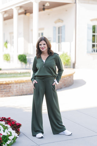 Woman posing in a stylish olive green two-piece loungewear set featuring a long-sleeve half-zip pullover top and matching high-waisted wide-leg pants. The bottoms showcase functional side pockets and front seam details that create an elevated athleisure look. Styled for a comfortable, sporty-chic aesthetic with crisp white athletic sneakers, photographed outdoors on a paved walkway in front of a classic building with white structural columns.