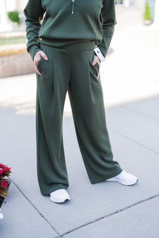 A close-up view focusing on the tailored details of the olive green wide-leg pants. The image highlights the deep functional side pockets, with the model's hands resting comfortably inside, and the structural front seams that add a refined, trouser-like touch to the relaxed fabric. The wide hem drapes perfectly over white athletic sneakers with black soles, emphasizing a casual, on-the-go style.