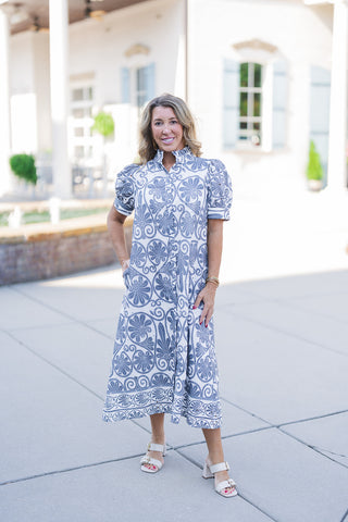 Woman posing outdoors in the Blue Botanical Scroll Midi Dress, a lightweight button-front piece featuring a striking large-scale blue scroll and floral pattern on a crisp white background. The dress features a ruffled mock neckline, short puff sleeves with circular eyelet cutouts, and functional side pockets. Styled with cream double-strap block-heel sandals, standing in front of an elegant white building with classical columns and light blue shutters.