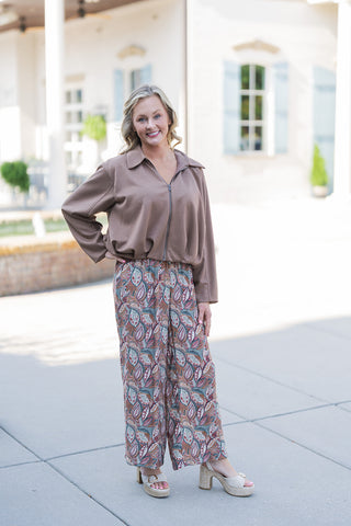 Woman posing in a casual yet elevated outfit featuring a solid mocha brown long-sleeve top with a classic collar, front zipper, and a gathered blouson hem. The top is paired with flowing wide-leg pants showcasing an intricate, earthy botanical paisley pattern in shades of teal, rust, cream, and brown. The look is styled for a relaxed, daytime aesthetic with woven beige espadrille platform sandals, photographed outdoors in front of a building with white structural columns.