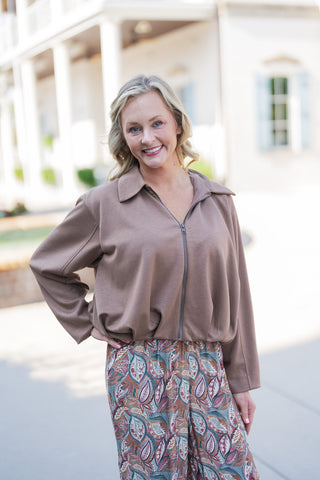A mid-shot portrait view highlighting the details of the solid mocha brown collared top. The image focuses on the functional dark metal front zipper pulled up to the collarbone and the soft, relaxed-fit fabric that gathers neatly at the elastic waist. This creates a flattering drape over the high-waisted, earth-toned botanical patterned trousers, offering a comfortable yet structured silhouette.