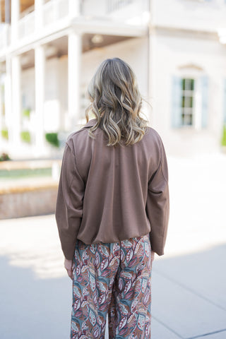 Back view of the outfit showcasing the clean, uninterrupted drape of the solid brown long-sleeve jacket and the breezy, full movement of the wide-leg patterned pants. The image highlights the relaxed fit across the shoulders and the gentle cinching at the waistline, emphasizing an effortless, transitional seasonal style perfect for everyday wear.